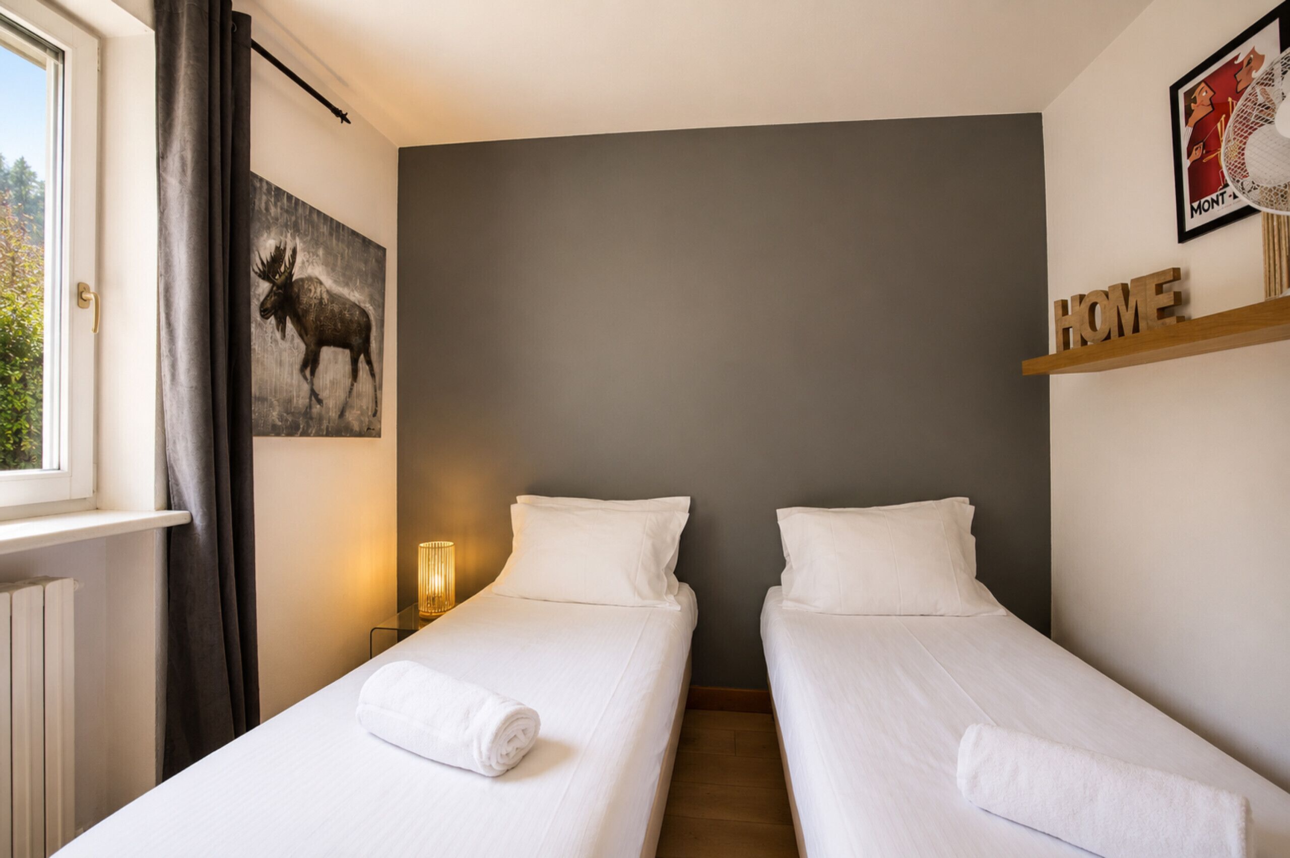 A hotel room with two beds, a moose painting, a 'HOME' sign, and a window with curtains.