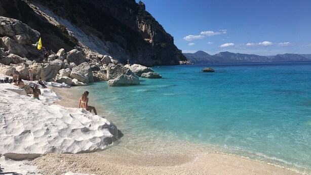 Une plage à l’eau turquoise et limpide, avec un terrain rocheux et un drapeau flottant au sommet d’une falaise.