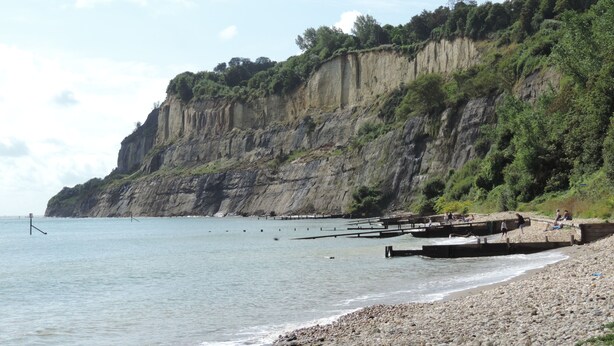 En stenet strand med en stejl klippe og en moler, der strækker sig ud i vandet.