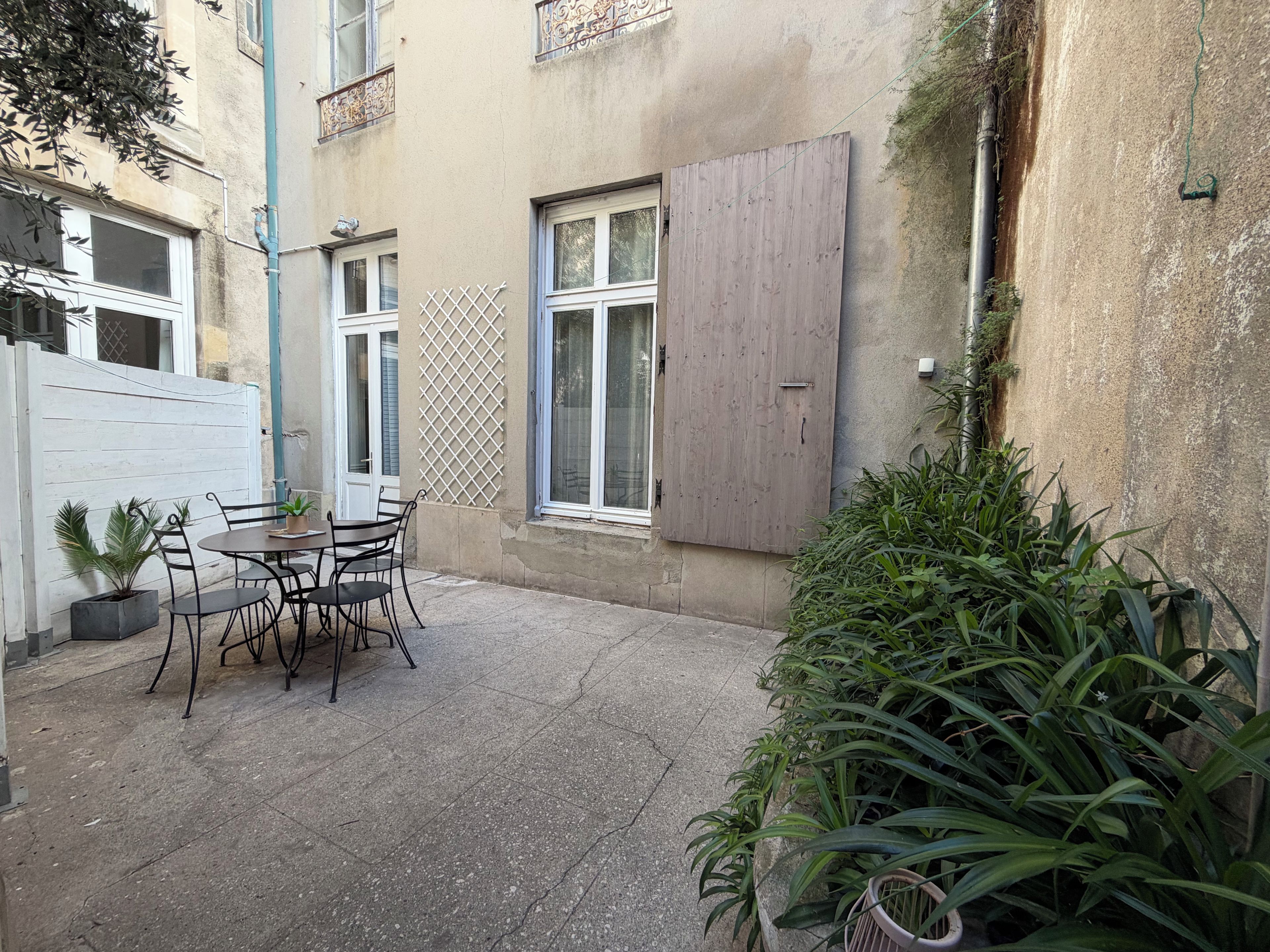 An outdoor patio with a table and chairs, a potted plant, and a wooden shuttered window.