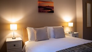 Een netjes opgemaakt bed met twee kussens, twee bedlampjes, een nachtkastje met boeken en een schilderij van een zonsondergang aan de muur.