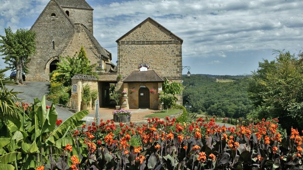 Una iglesia de piedra con un campanario y un edificio más pequeño, rodeados de exuberante follaje y flores coloridas.