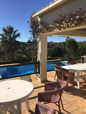 Zona junto a la piscina con sillas de mimbre y una mesa, con vista a una piscina y exuberante vegetación.