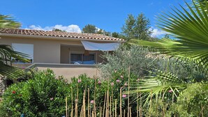 Una casa con techo de tejas, un porche cubierto y abundante vegetación, incluyendo palmeras y plantas con flores.