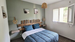 Un dormitorio con cabecera de madera, cama con cubrecama azul estampado, ventana con cortinas blancas y cuadros colgados en la pared.