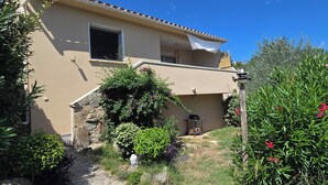 Una casa de dos pisos con fachada color beige, un muro de piedra y un jardín con diversas plantas y flores.