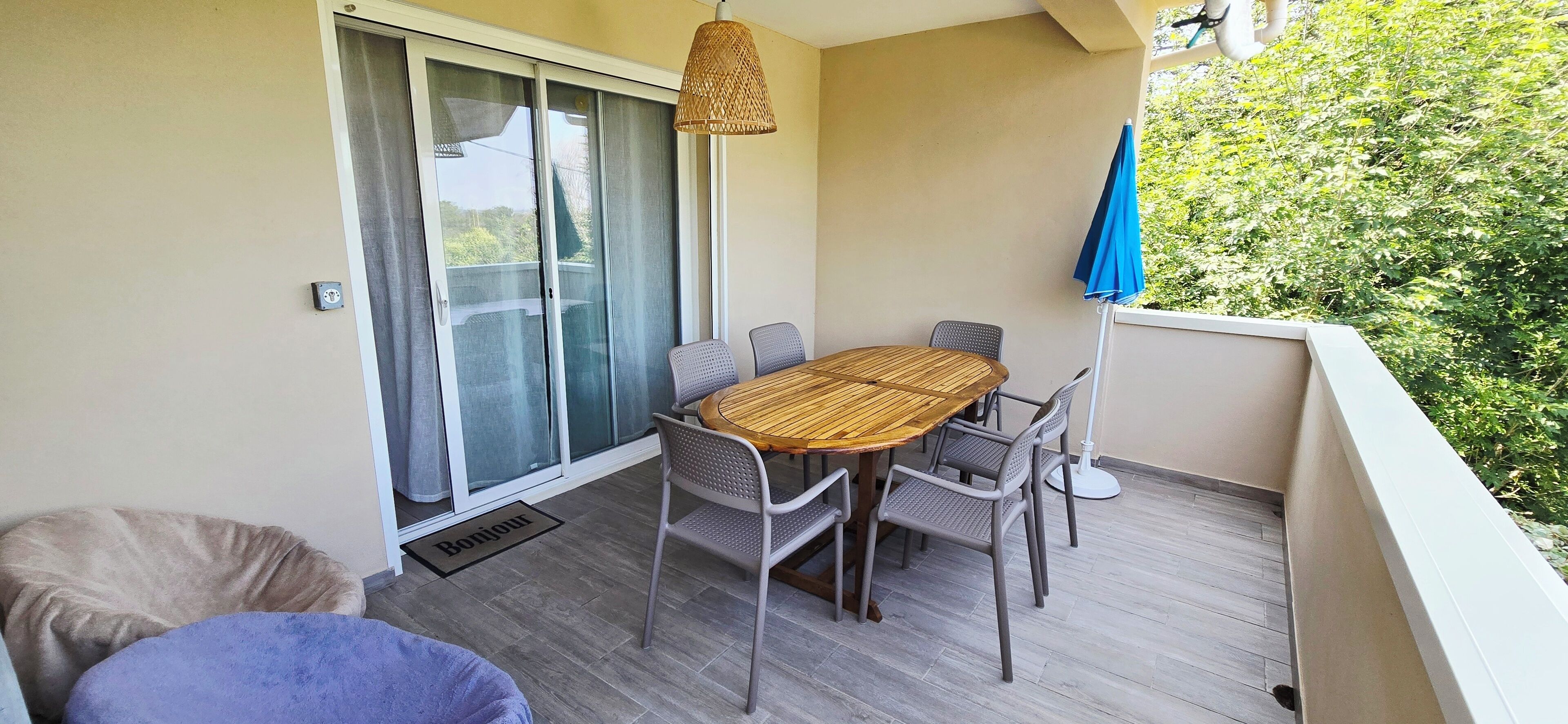 A balcony with a wooden table and chairs, a wicker lamp, and a blue umbrella.