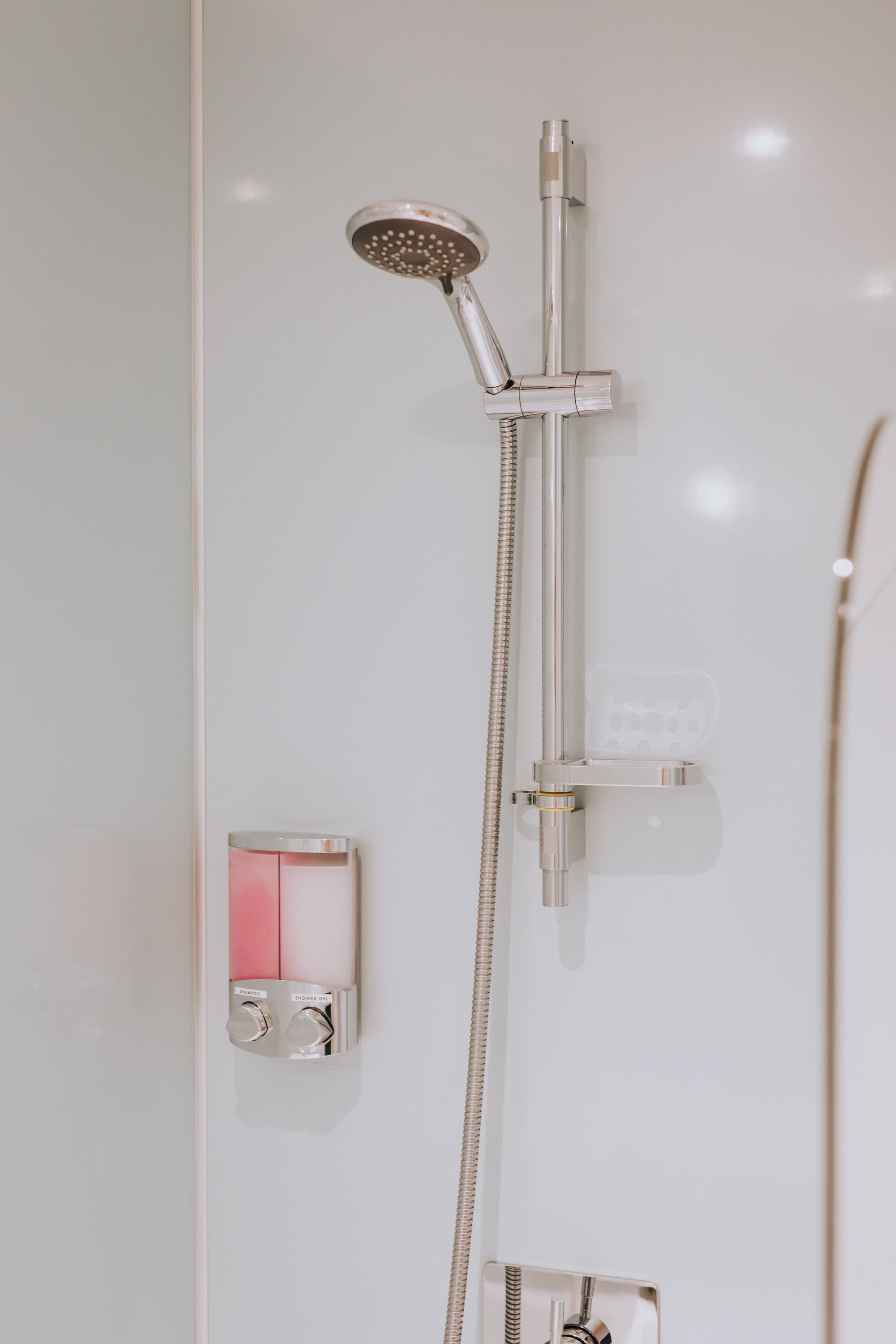 Eine moderne Dusche mit Handbrause und einer wandmontierten Bedienkonsole mit den Aufschriften 'Shampoo' und 'Duschgel'.