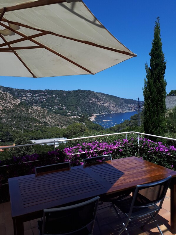 A terrace with a table and chairs, overlooking a coastal landscape with hills and a body of water.