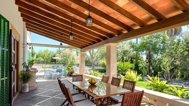 Ein überdachter Außenbereich mit Tisch und Stühlen, umgeben von Pflanzen und einem Pool.