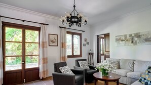Ein Wohnzimmer mit einer Couch, zwei Sesseln, einem Couchtisch und einer Chandelier. Große Fenster mit Vorhängen, eine Tür und ein Spiegel an der Wand sind ebenfalls vorhanden.