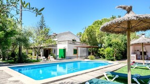 Ein Swimmingpool mit Liegestühlen und einem Reetdach-Sonnenschirm, umgeben von viel Grün und einem Gebäude mit einer grünen Tür.