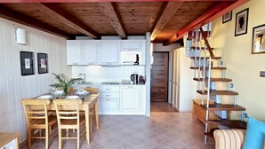 Une cuisine compacte avec des meubles blancs, un îlot central et un coin repas doté de chaises en bois. Un escalier avec une rampe en bois mène à l’étage supérieur.
