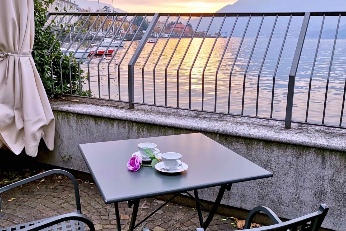 Une table avec deux tasses blanches et une rose rose, placée sur un balcon donnant sur une étendue d’eau, avec des bateaux et des bâtiments en arrière-plan.