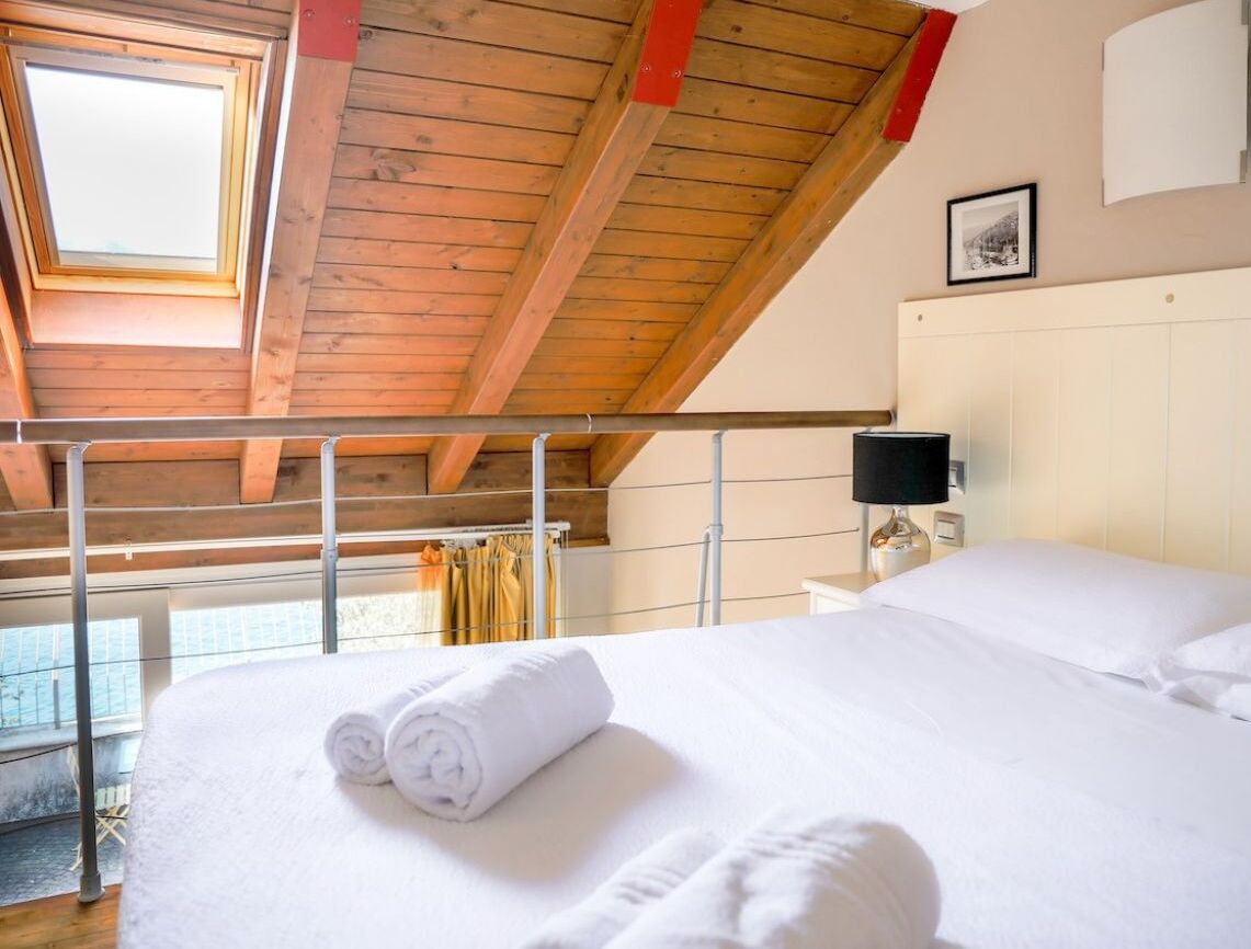 A bedroom with a wooden ceiling, a bed with white linens, a bedside table with a lamp, and a window with curtains.