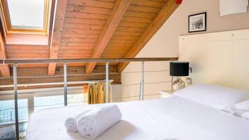 A bedroom with a wooden ceiling, a bed with white linens, a bedside table with a lamp, and a window with curtains.