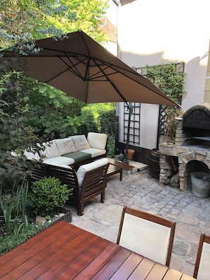 An outdoor patio with a wooden table and chairs, a cushioned sofa, and a stone oven.