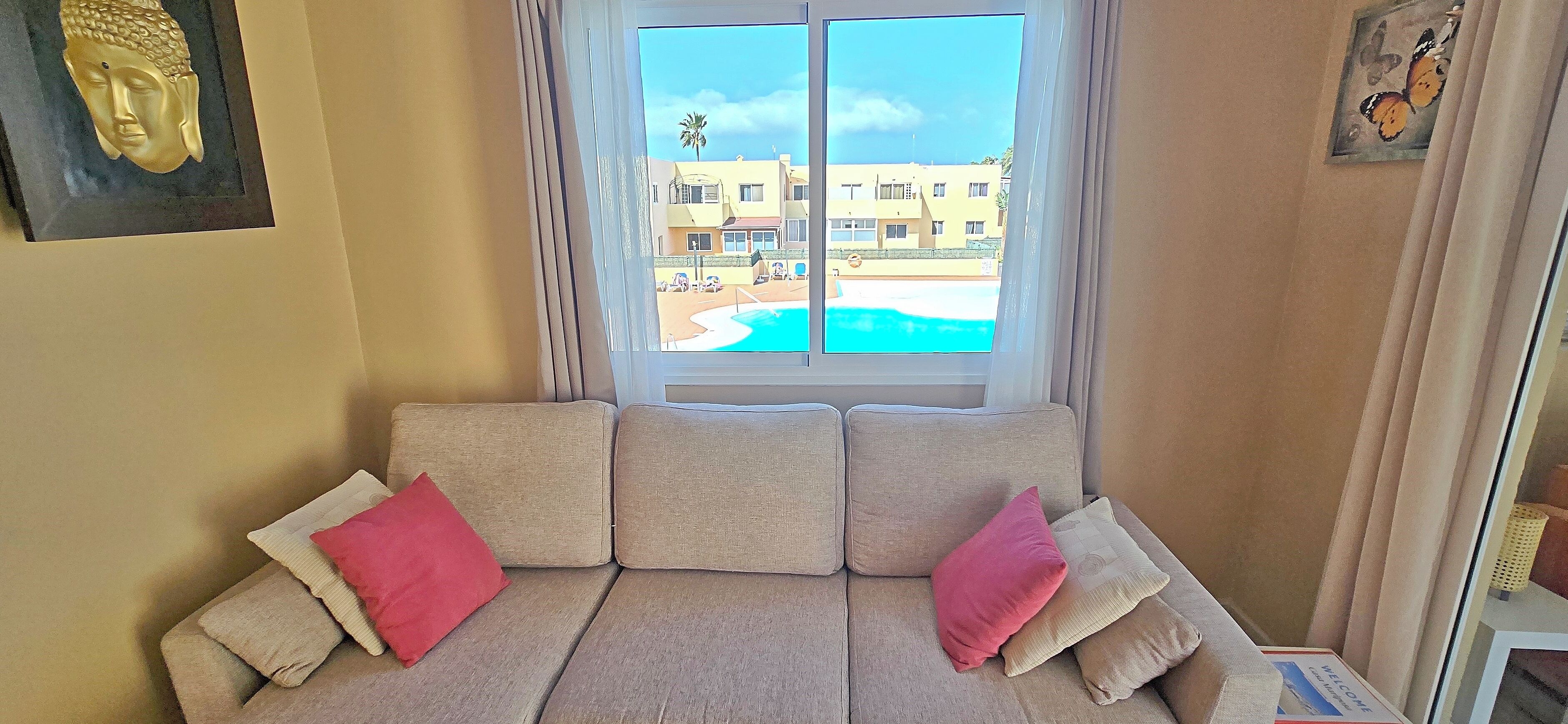 Ein beiges Sofa mit rosafarbenen und weißen Kissen, eine Buddha-Kopf-Skulptur an der Wand und Blick auf einen Pool und Gebäude durch ein Fenster.