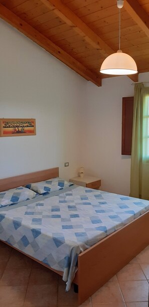 A bedroom with a wooden ceiling, a bed with a patterned bedspread, a bedside table with a lamp, and a painting on the wall.