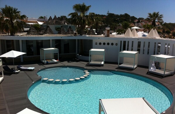 A swimming pool with lounge chairs and umbrellas, surrounded by palm trees and a building with a modern design.