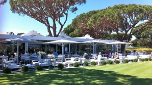 An outdoor seating area with white tables and umbrellas, surrounded by trees and a well-maintained lawn.
