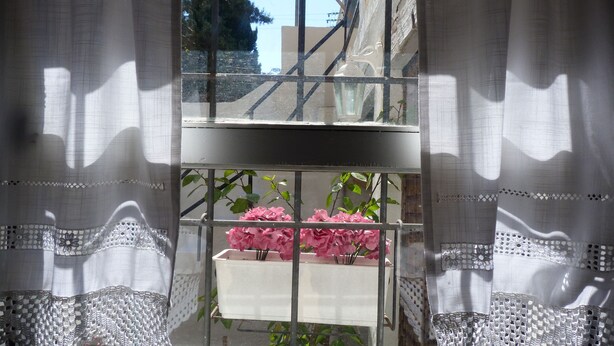 A window with a view of a balcony, a white flower pot with pink flowers, and a metal railing.