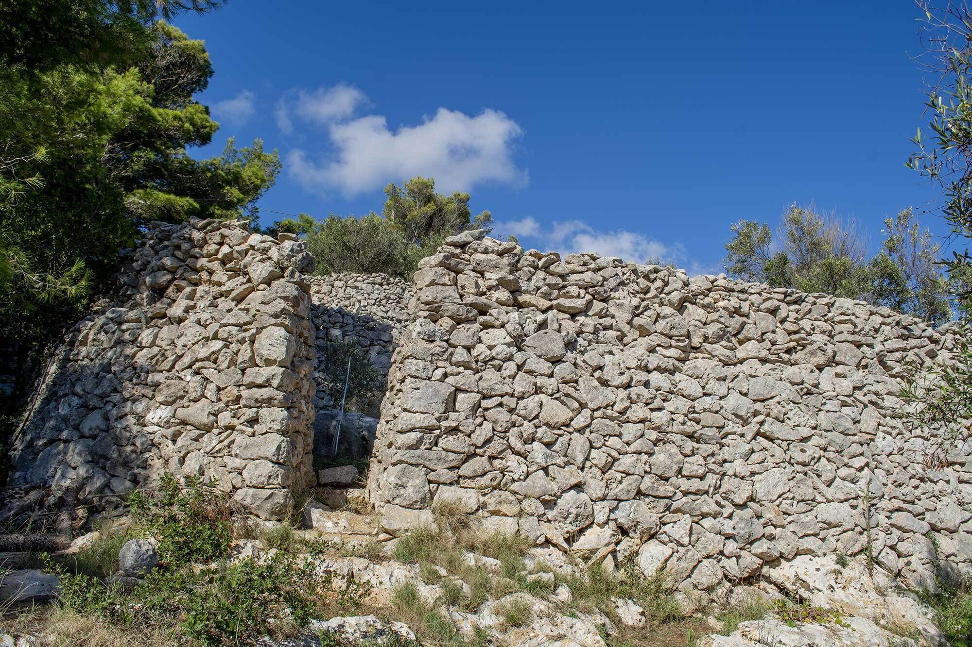 Un mur de pierre percé d’une étroite ouverture, entouré de végétation naturelle et d’arbres.