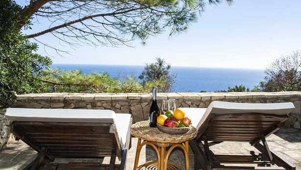 A seaside terrace with two wooden lounge chairs, a wicker table with a fruit bowl, a bottle, and glasses.