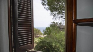 A view through a window with wooden shutters, overlooking a green landscape with trees and a distant view of the sea.