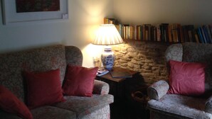 A cozy living room with two patterned armchairs, a small table with a lamp, a bookshelf, and a painting on the wall.