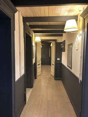 Een gang met houten vloeren, witte wanden en zwarte deuren. Twee wandlampen zorgen voor verlichting.