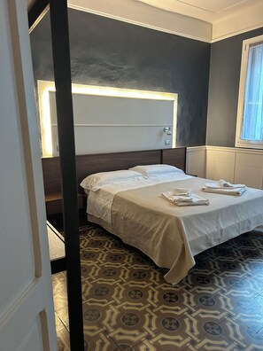Een hotelkamer met een bed, hoofdbord, nachtkastje, lamp en vloertegels met een patroon.
