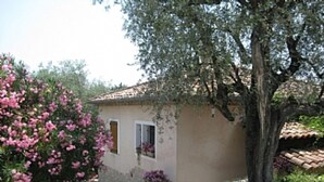 Un petit bâtiment avec un toit en tuiles, une fenêtre et une porte. Un arbre se trouve devant le bâtiment et un buisson aux fleurs roses est situé sur la gauche.