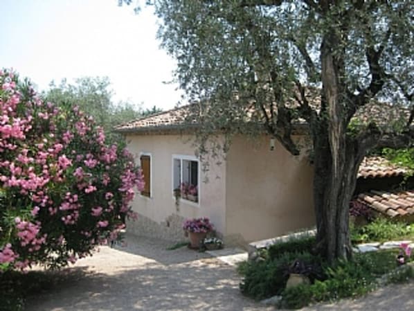 Un petit bâtiment avec un toit en tuiles, une fenêtre et une porte. Un arbre se trouve devant le bâtiment et un buisson aux fleurs roses est situé sur la gauche.