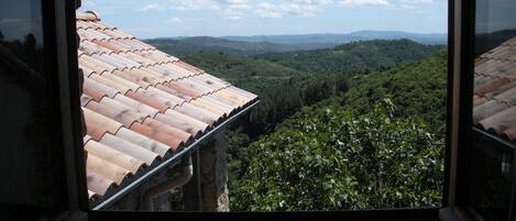Une vue depuis une fenêtre, sur un toit recouvert de tuiles, donnant sur une vallée verdoyante et des collines lointaines.