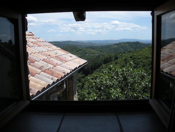 A view from a window with a tiled roof, overlooking a valley with greenery and distant hills.