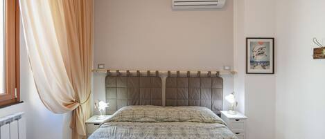 Un dormitorio con una cama, dos mesitas de noche, aire acondicionado, una ventana con cortinas y un cuadro en la pared.