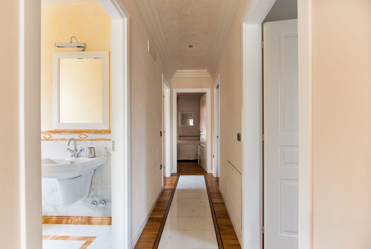 Un corridoio con pavimento in legno, pareti bianche e, a sinistra, un bagno con lavandino e specchio.