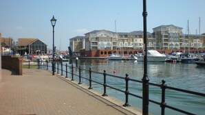 A waterfront promenade with a brick walkway, metal railings, and a row of buildings with balconies overlooking the water. - Fabulously Located Marina Apartment - Superb Waterside Views from every window. (Eastbourne)