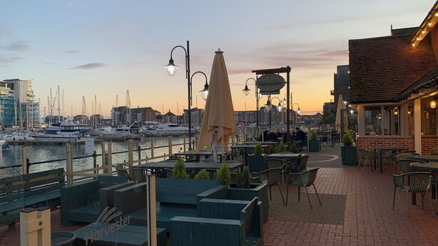 A waterfront patio with outdoor seating, a view of the marina, and a building with a sign reading "Harvester". - Fabulously Located Marina Apartment - Superb Waterside Views from every window. (Eastbourne)