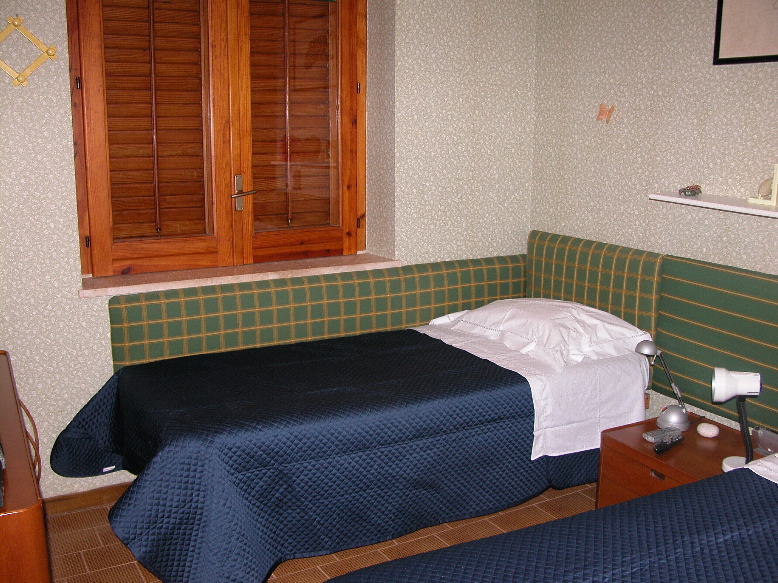 A small room with two beds, a window with wooden shutters, a green checkered bench, and a small table with a lamp and remote control.