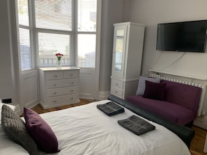 Ein Schlafzimmer mit Bett, Kommode, Fernseher und einem Fenster mit Jalousien.