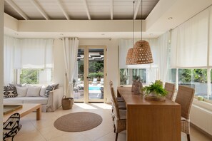 A modern living room with a sofa, dining table, and chairs, a large window with curtains, and a view of an outdoor pool. - Levantes Luxury Home - Nature Inspired, Pool & BBQ (Rethymno)