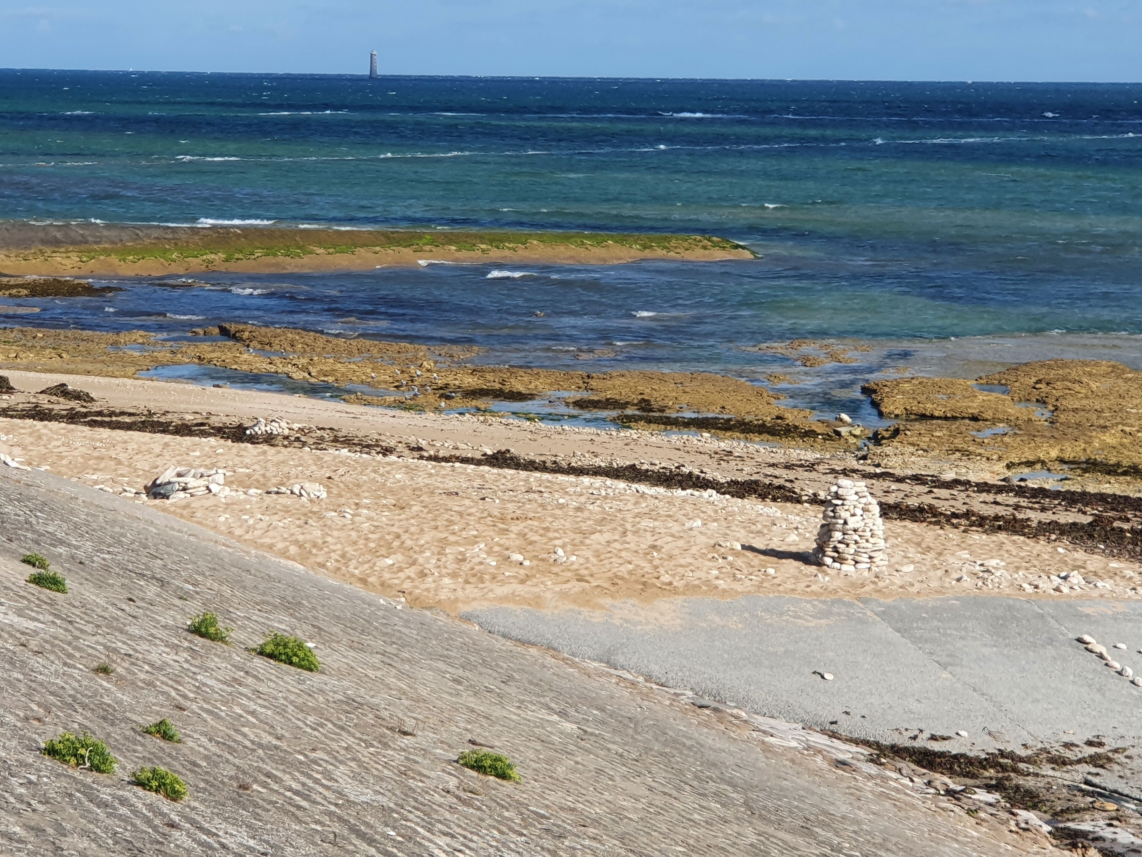 Ein felsiger Strand mit klarem, blauem Wasser und einem fernen Leuchtturm.