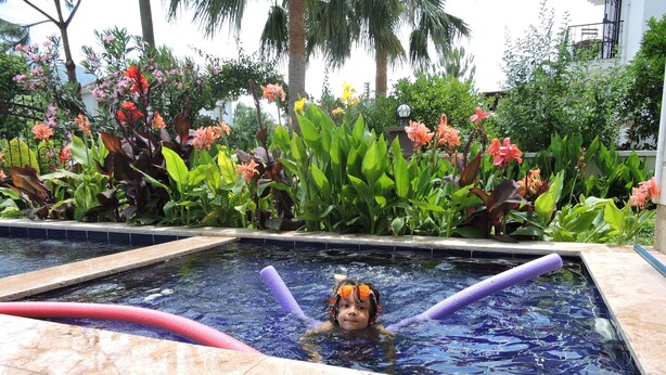 Criança em piscina com boia de segurança, rodeada por vegetação tropical exuberante.