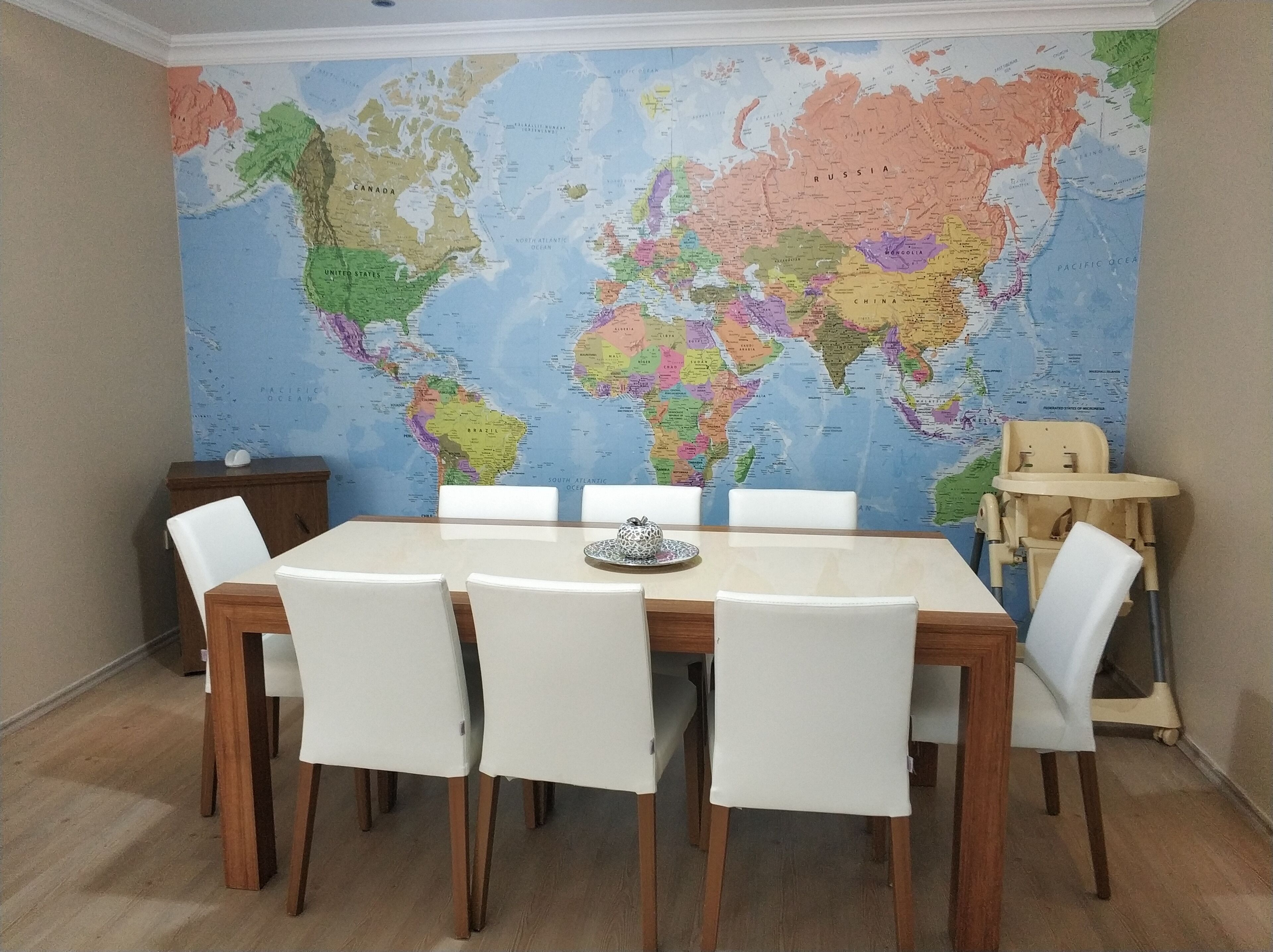 Un comedor con un gran mapa mundial en la pared, una mesa de madera y sillas blancas.