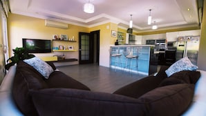 Sala de estar com sofá, televisão e área de cozinha equipada com geladeira, micro-ondas e banquetas.