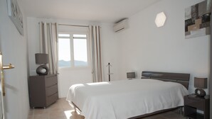 A bedroom with a large bed, a dresser, a bedside table with a lamp, a window with curtains, and an air conditioning unit. - VILLA 4 ROOMS 3 BATHROOMS WITH  HEATED POOL  500M SEA VIEW / MOUNTAIN (Zonza)