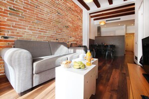 Uma sala de estar moderna com um sofá cinzento, uma mesa de centro branca com frutas e uma garrafa, e uma parede de tijolos.