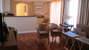 Une chambre d’hôtel avec un plancher en bois, un canapé, deux fauteuils en osier, une petite table avec un vase et un coin cuisine comprenant un comptoir et des placards.